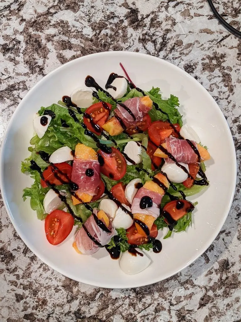 green salad with peaches, tomatoes, and mozzarella balls with balsamic glaze drizzled over ingredients in white bowl